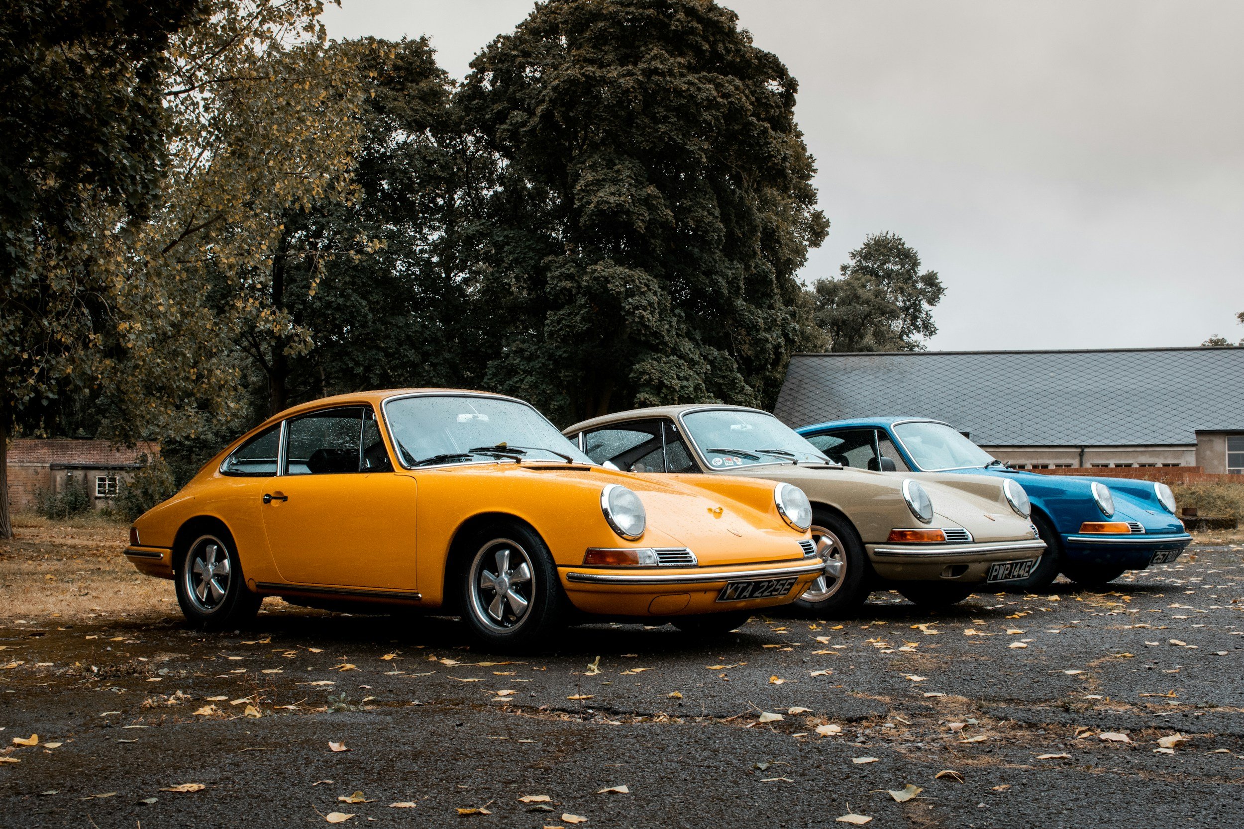 Three vintage Porsche 911 cars in yellow, beige, and blue parked outdoors on a leaf-strewn surface in front of trees and a building.