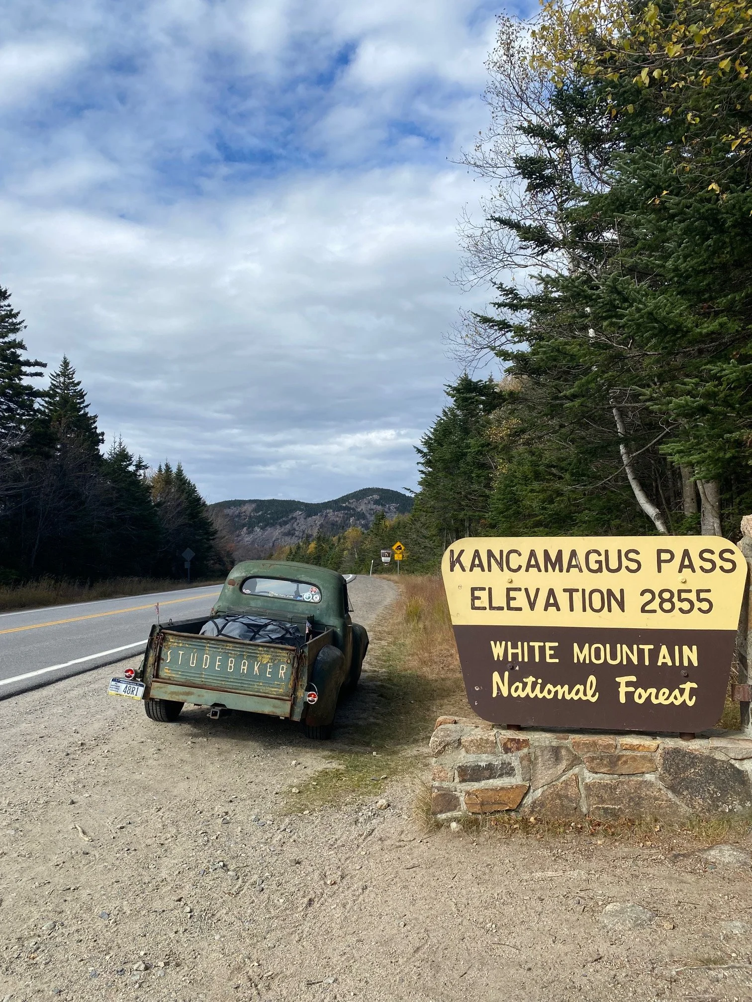 A rural mountain roadside scene with a vintage blue pickup truck parked beside a sign that reads "Kancamagus Pass, Elevation 2855, White Mountain National Forest." There are trees and mountains in the background under a cloudy sky.