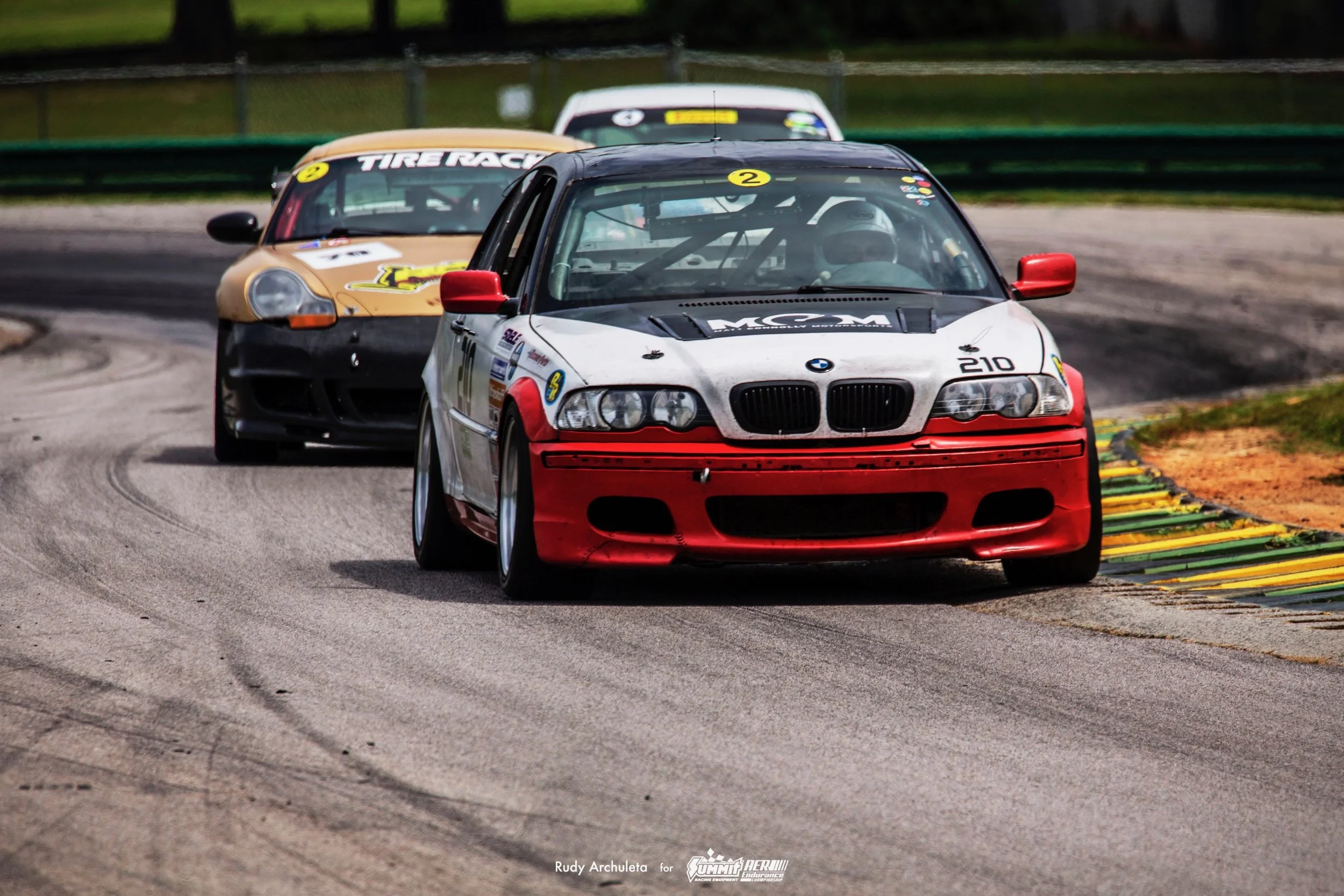 Three race cars on a track, maneuvering a turn. The front car is a red and white BMW with the number 210, followed by a beige and black car, and a white car in the background.