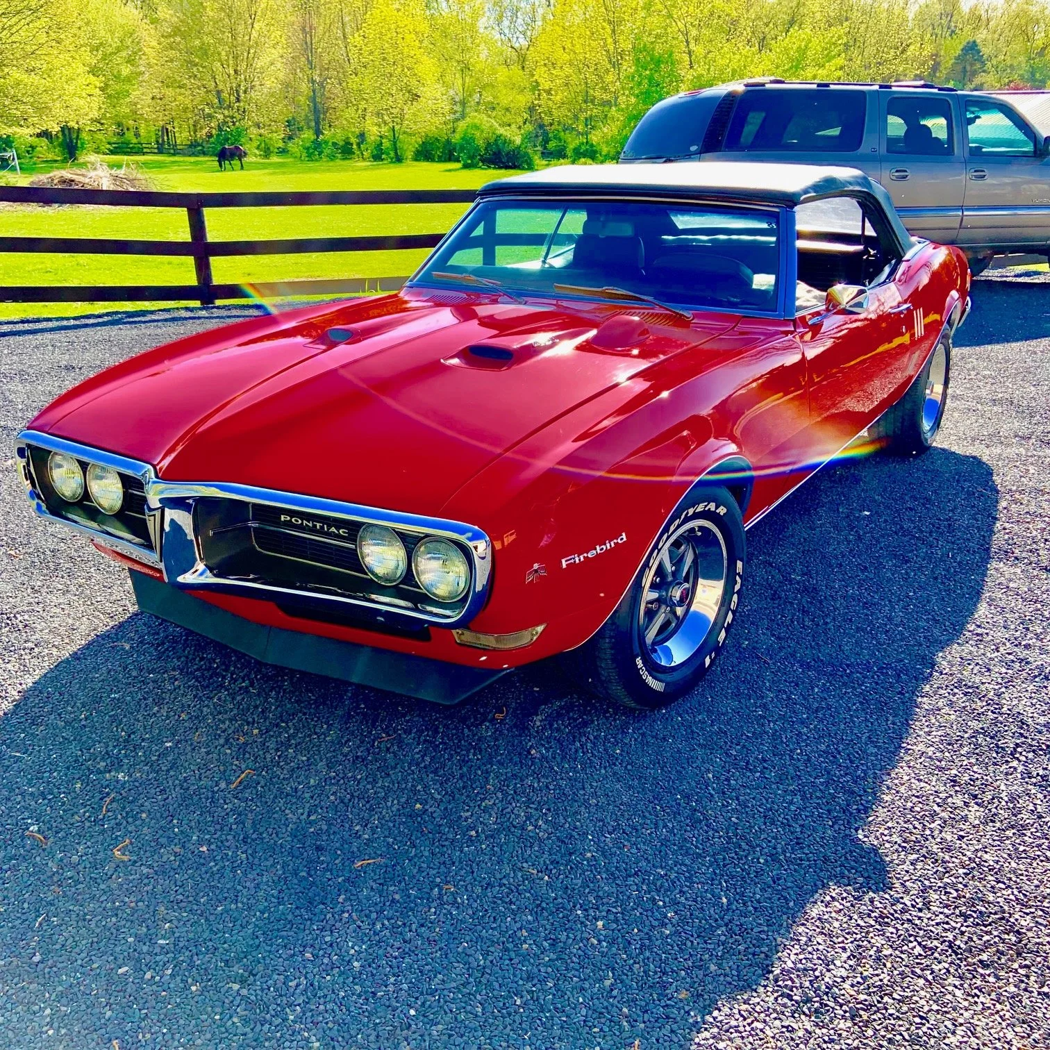 A classic red Pontiac Firebird convertible with a white top is parked on a driveway next to a gray SUV, with a green field and trees in the background.