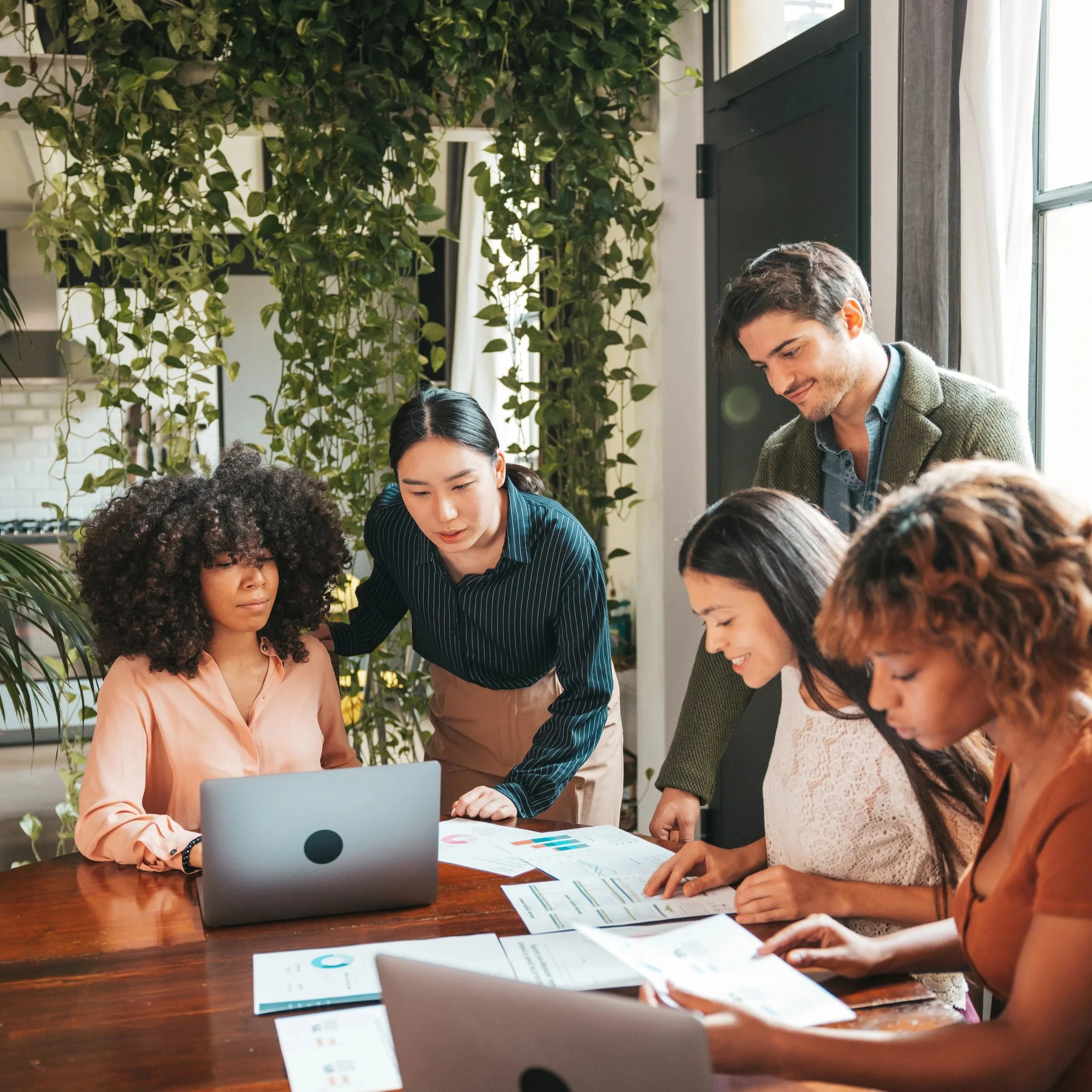 A diverse group of five people collaborating around a wooden table with laptops and documents, in a bright space with green plants.