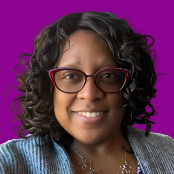 A woman with curly dark hair, wearing red glasses, smiling, against a purple background