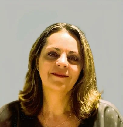 A woman with shoulder-length brown hair, smiling, wearing a black top with small dots, standing against a plain light background.