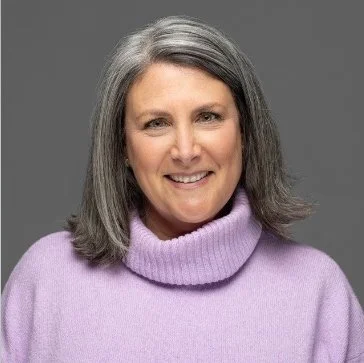 A woman with shoulder-length gray hair smiling, wearing a lavender turtleneck sweater against a gray background.