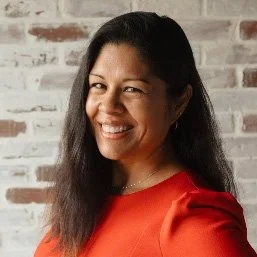 A woman with long dark hair smiling and winking, wearing a red top, standing in front of a brick wall background.