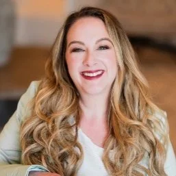 A smiling woman with long, wavy blonde hair, wearing a light-colored blazer and a white top.