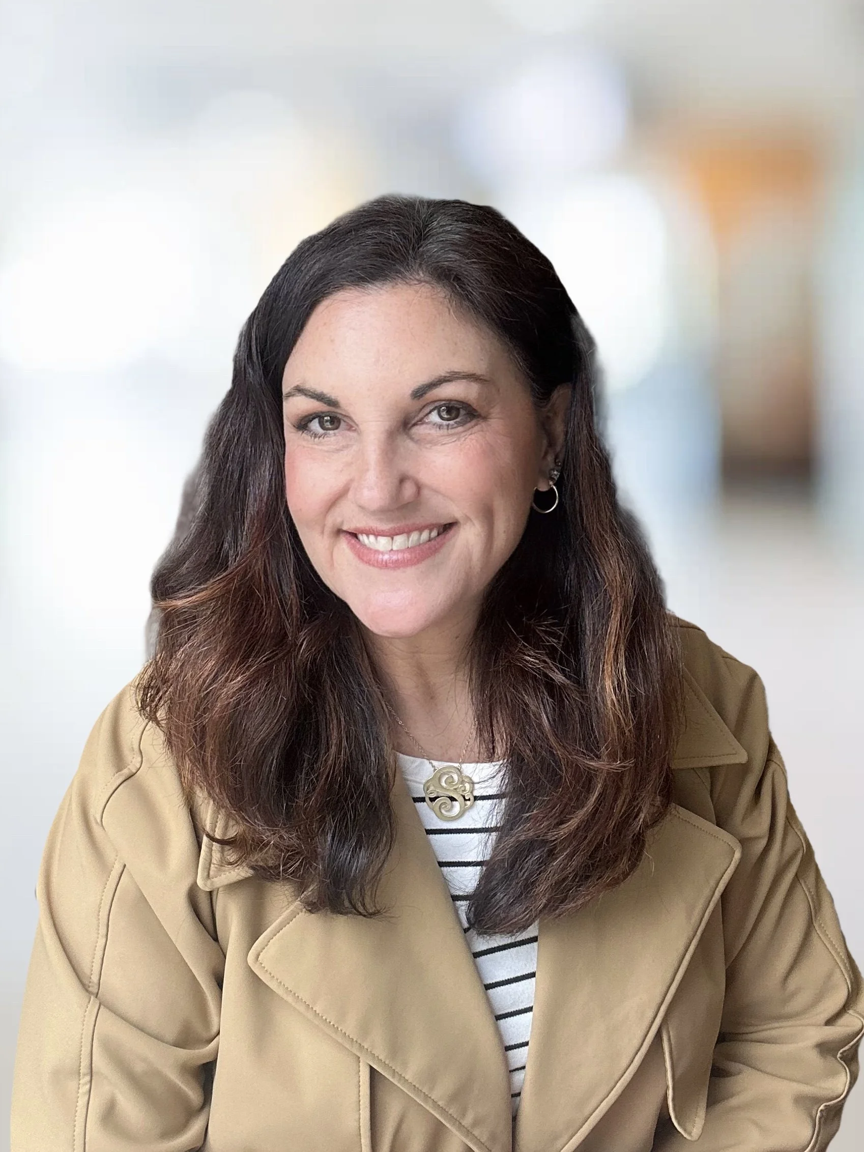 Suzi Tropiano with dark brown hair and a bright smile, wearing a tan jacket and striped top, in front of a blurred background.