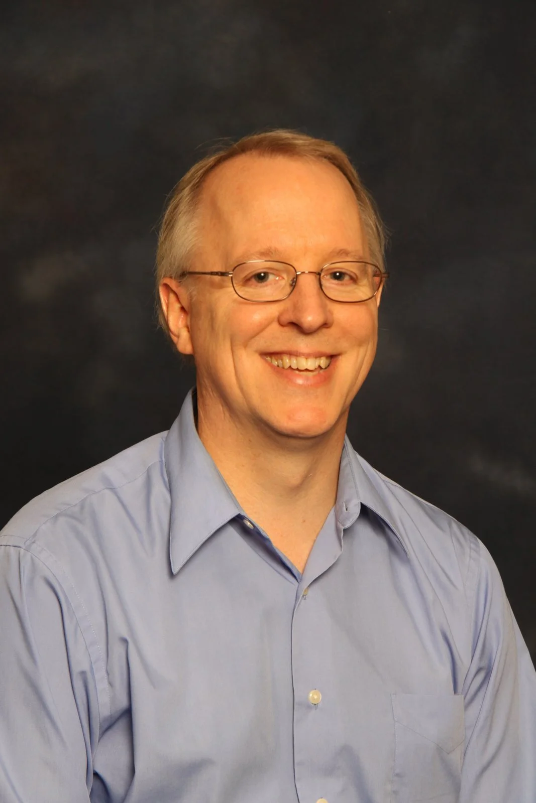 Eric Derby - a smiling man with glasses, wearing a light blue collared shirt, against a dark background.