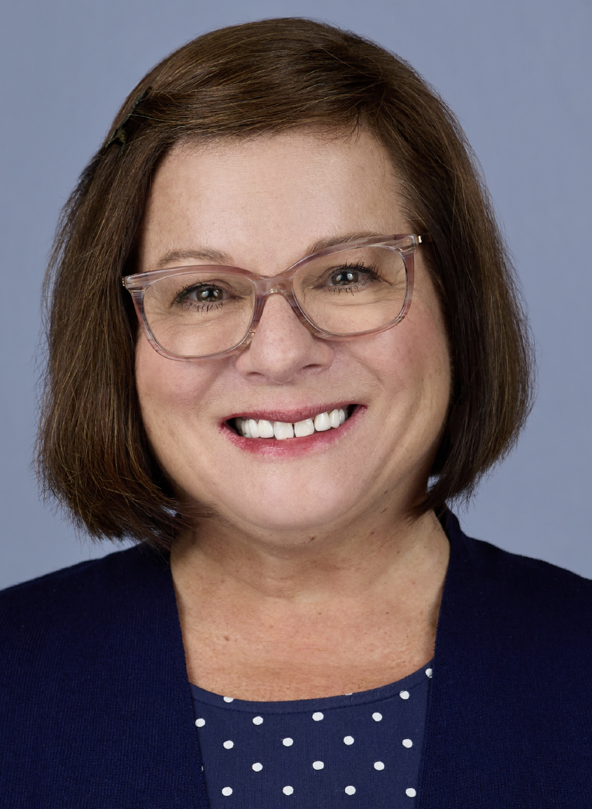 Patti Maenza with shoulder-length brown hair wearing glasses, a navy blazer, and a polka-dot shirt, smiling against a light blue background.