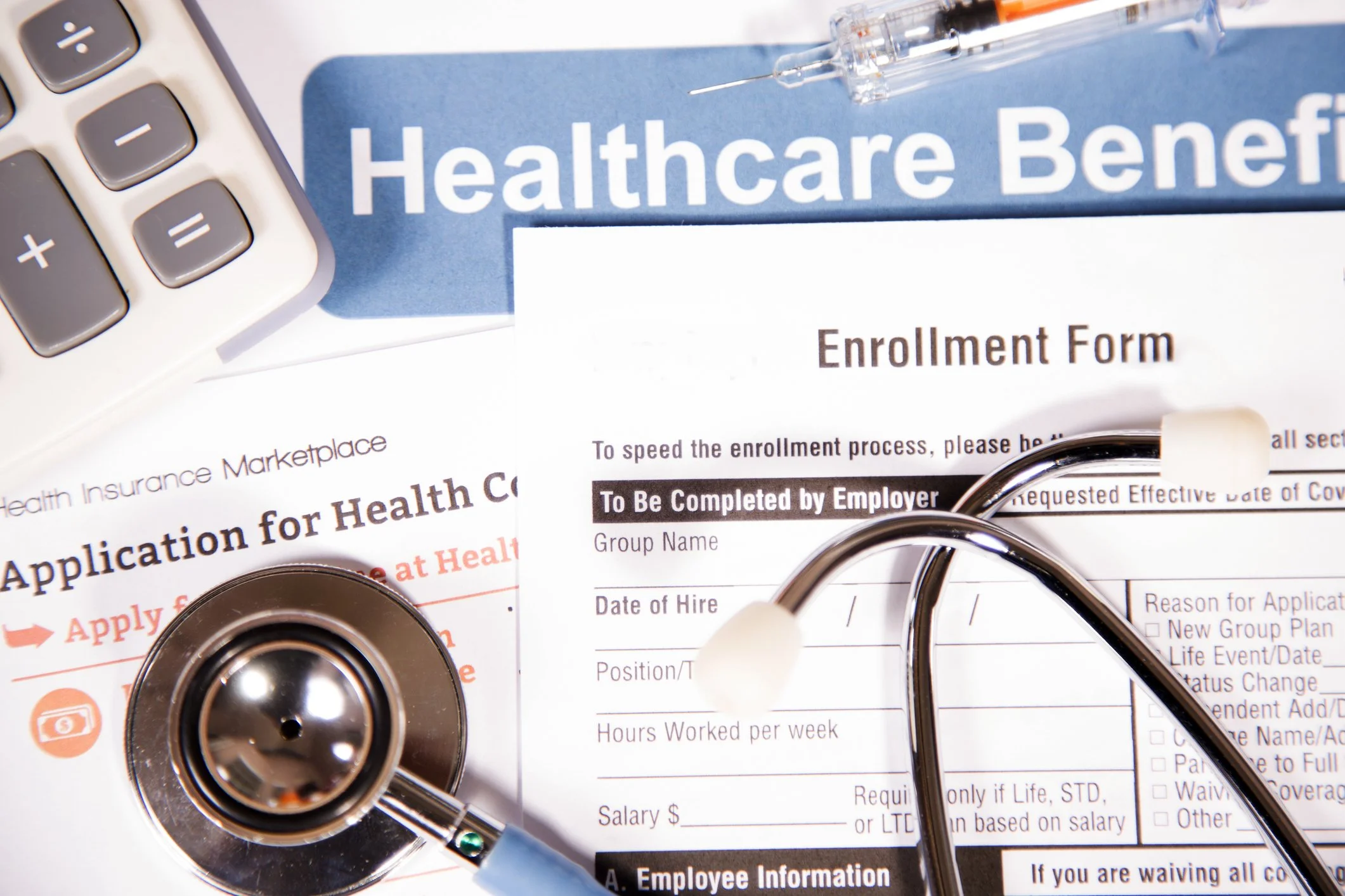 Stethoscope, insurance enrollment form, and a calculator on top of a document labeled 'Healthcare Benefits' and 'Application for Health Coverage'.
