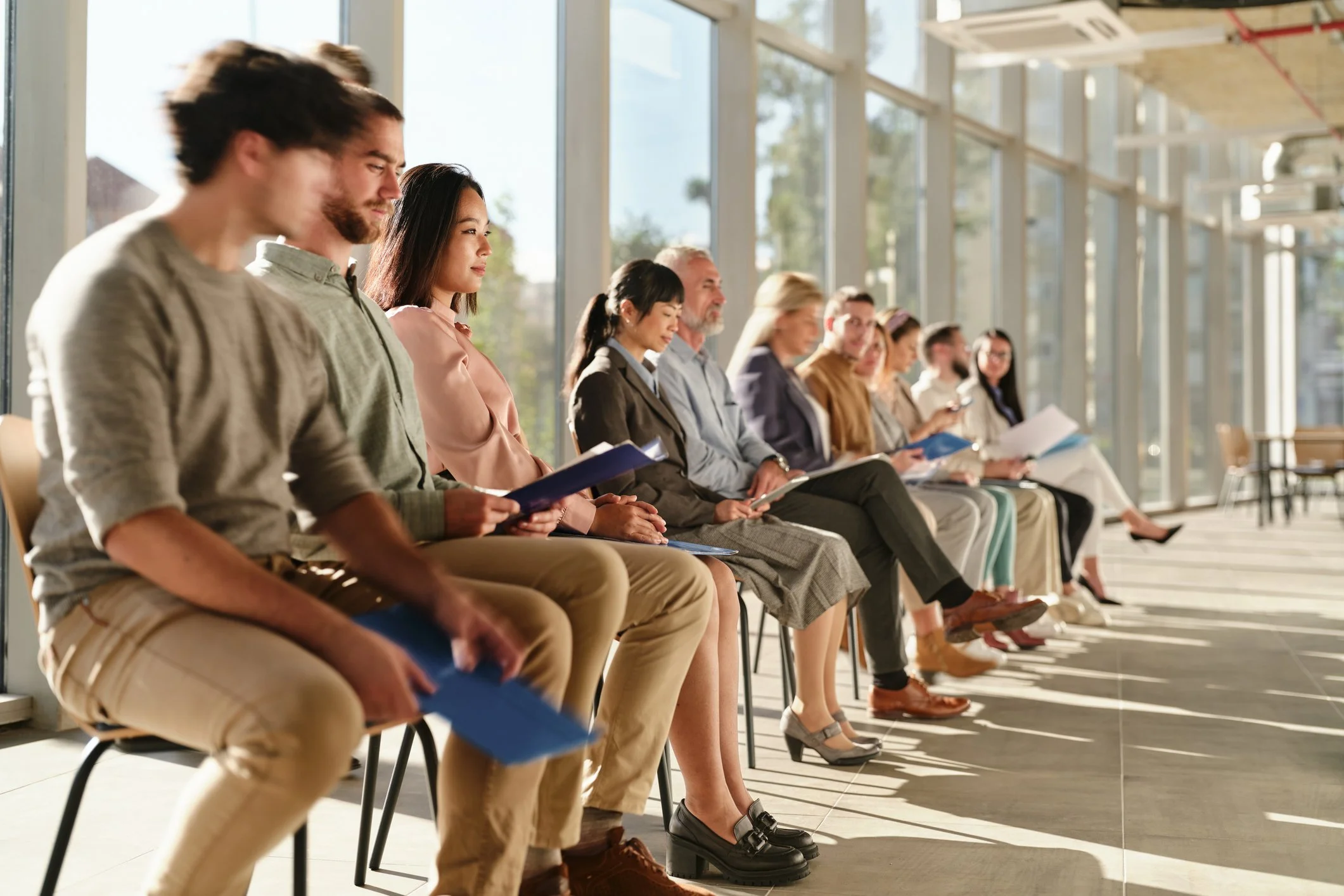 People sitting in a row at a waiting area, reading or waiting, with sunlight coming through large windows.