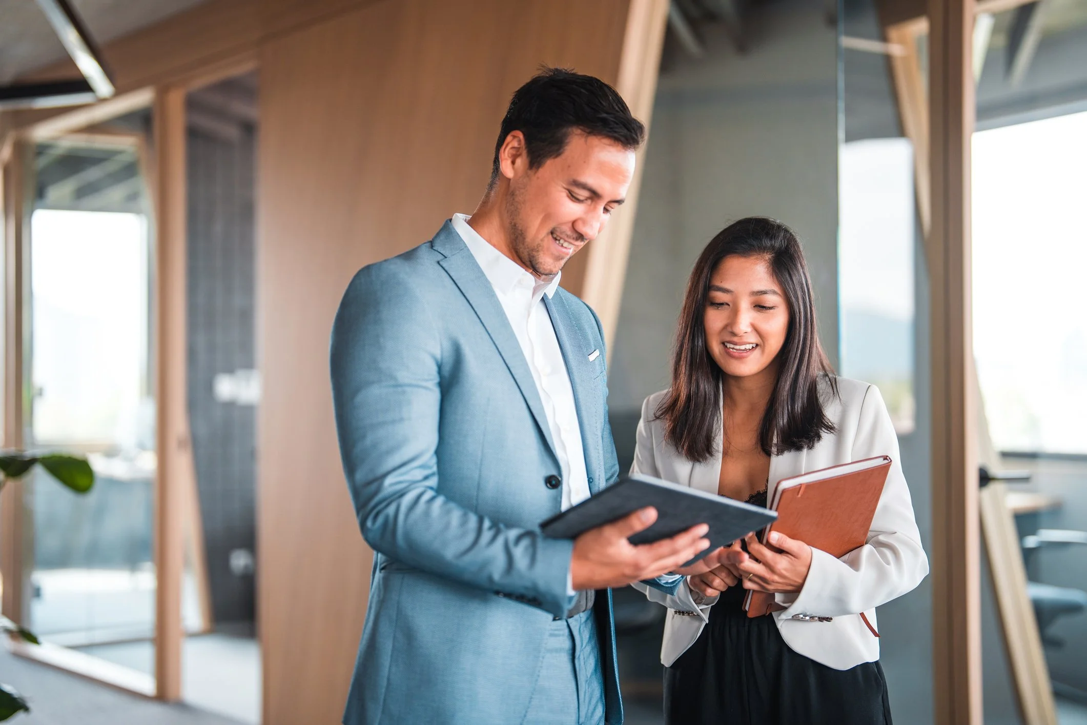 Two professionally dressed individuals, a man and a woman, are standing and looking at documents and a tablet together in a modern office space with large windows and wooden interior accents.