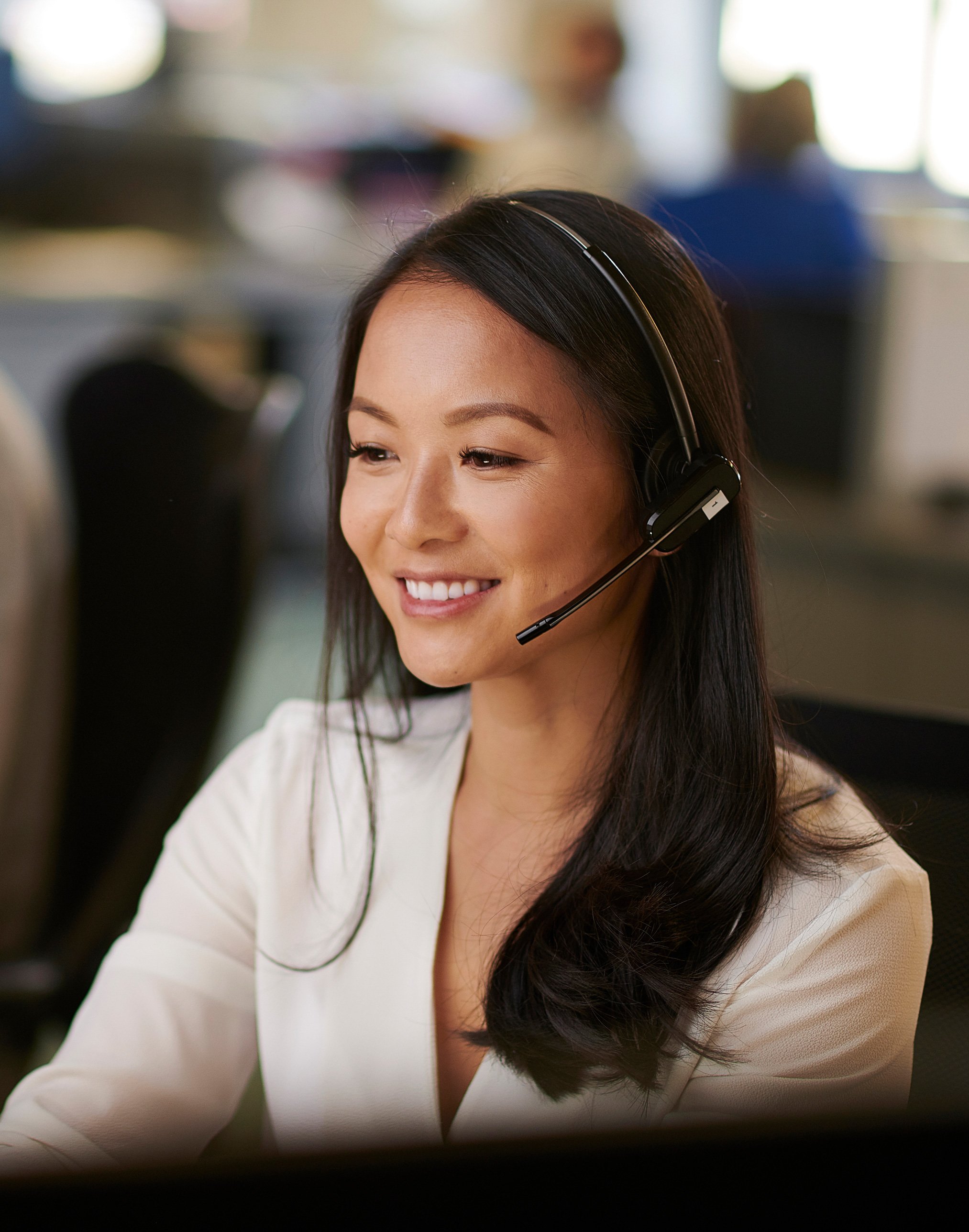 A woman with long dark hair wearing a headset, smiling while working at a computer in an office setting.