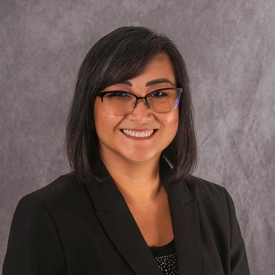 Founder, TU Jacobs, with shoulder-length black hair, wearing glasses and a black blazer, smiling against a neutral gray background.