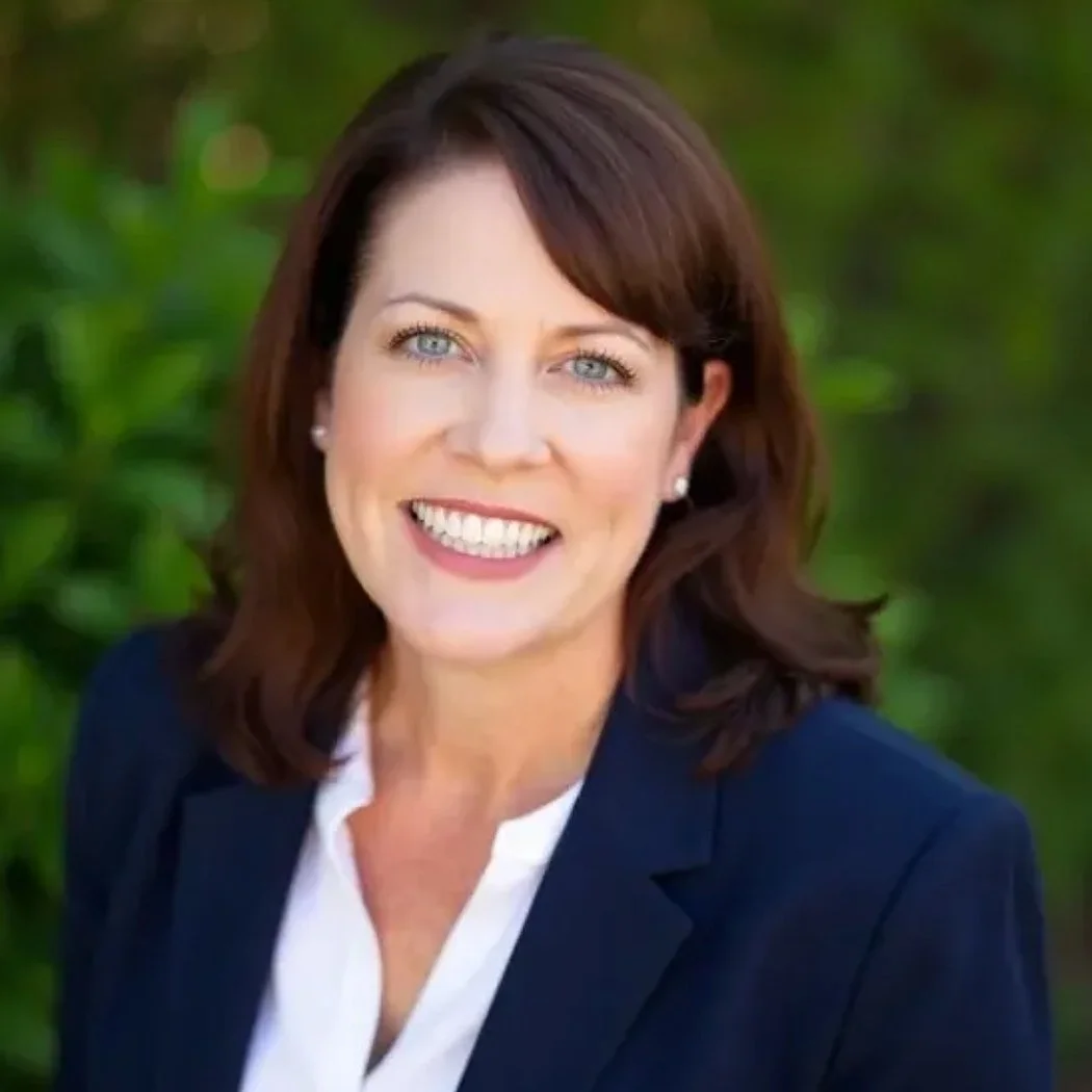 A smiling woman with shoulder-length brown hair, wearing a navy blazer and a white shirt, outdoors with green foliage in the background.