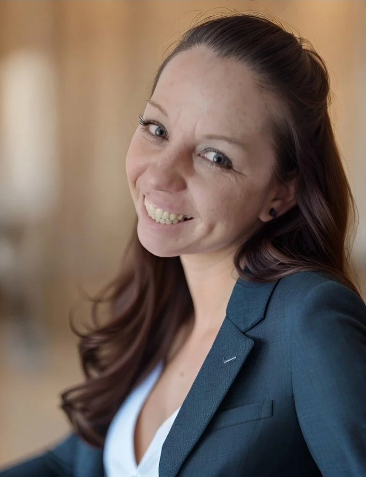 Smiling woman with long brown hair tied back, wearing a navy blazer and white top, indoors.