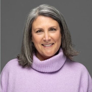 Smiling woman with shoulder-length gray hair wearing a lavender turtleneck sweater against a gray background.