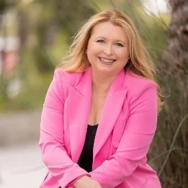 A woman with blonde hair wearing a pink blazer and a black top, smiling outdoors with greenery and trees in the background.