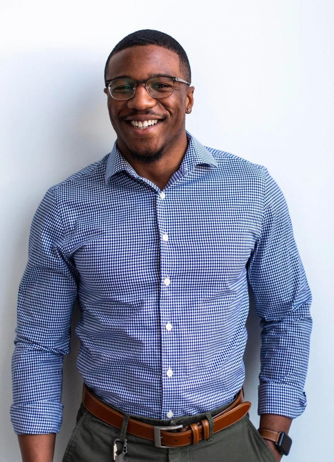 A smiling young man wearing glasses, a checkered blue and white shirt, dark pants, brown belt, and a smartwatch, standing in front of a plain white wall.