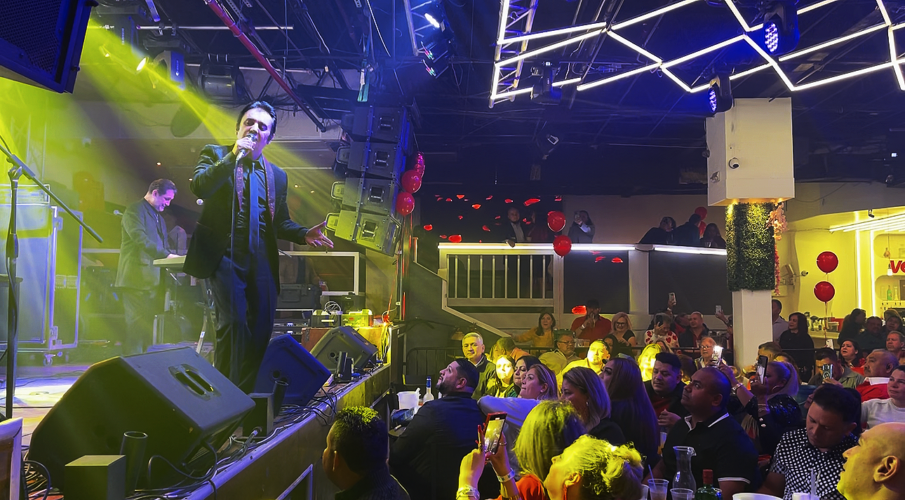 A singer performing on stage at a live music venue with a keyboardist in the background, audience members watching, some taking photos, colorful stage lights, and a lively atmosphere.