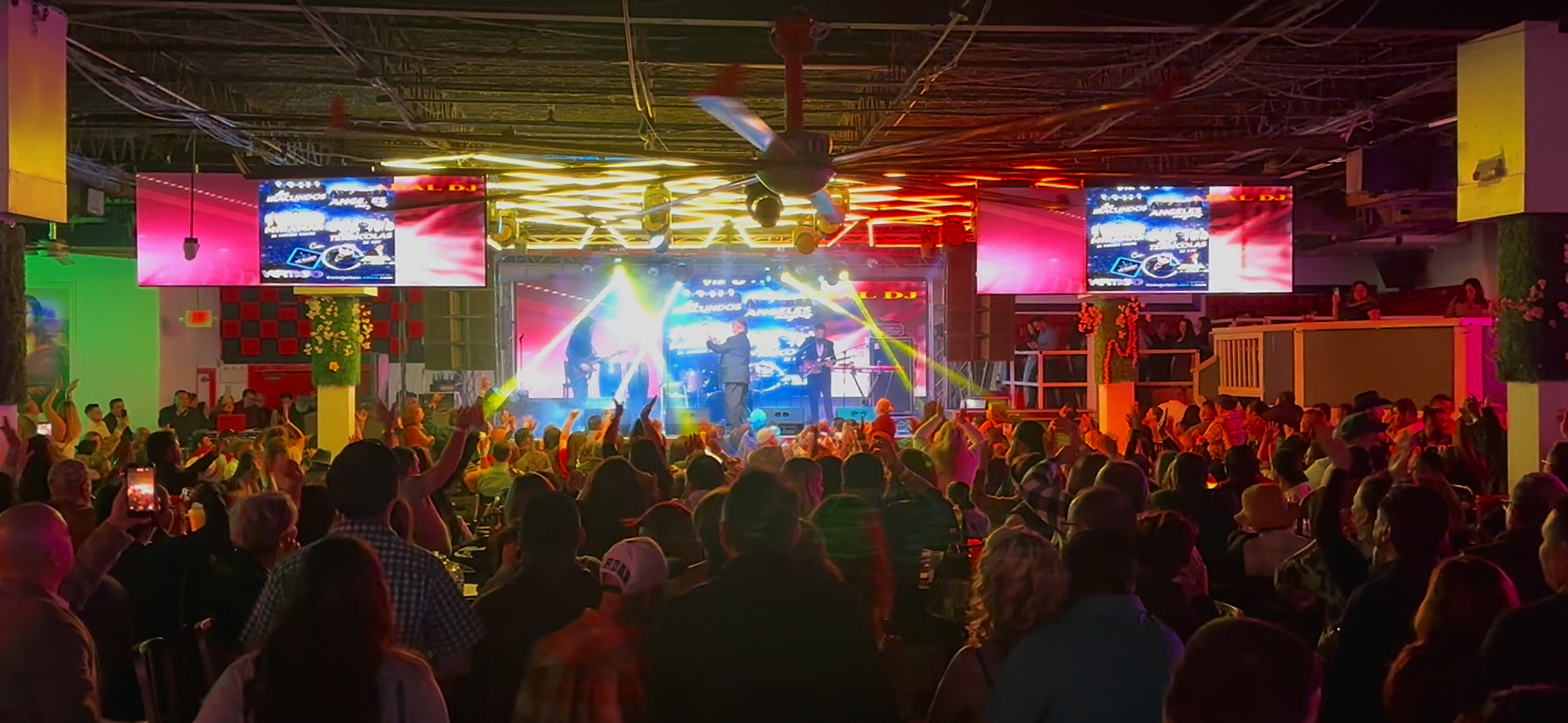 Crowd watching a live band perform on stage with colorful lighting and large screens in a lively indoor venue.