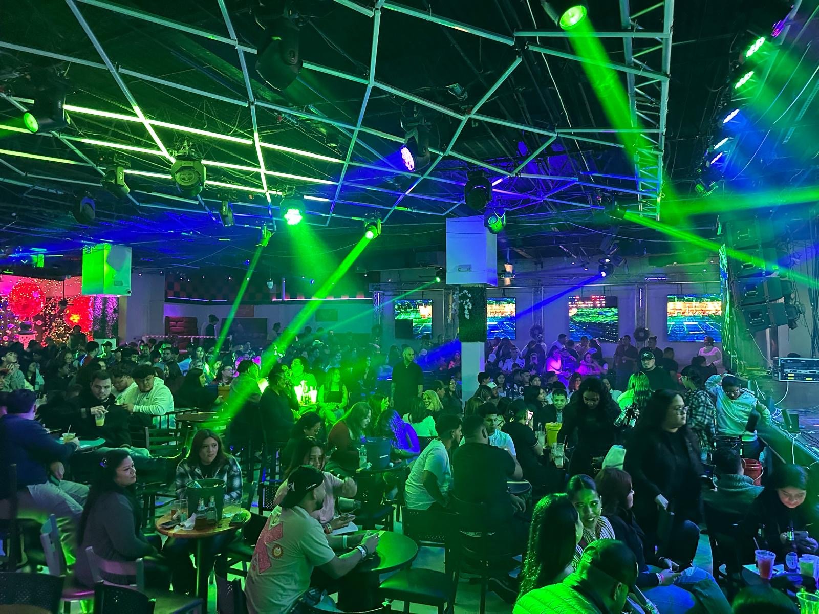A crowded indoor dance club bathed in green and blue laser lights, with people sitting at tables and watching a DJ performance.