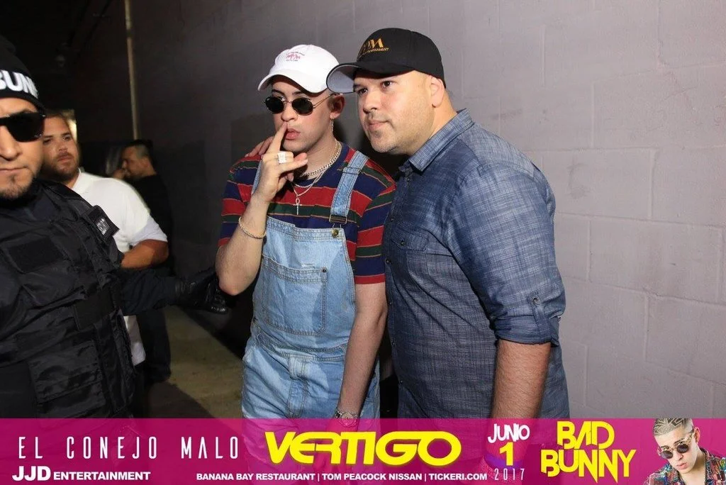 Two men standing against a brick wall, one wearing a black cap and a blue shirt, the other in a white cap, sunglasses, striped shirt, and overalls, with a security officer beside them.