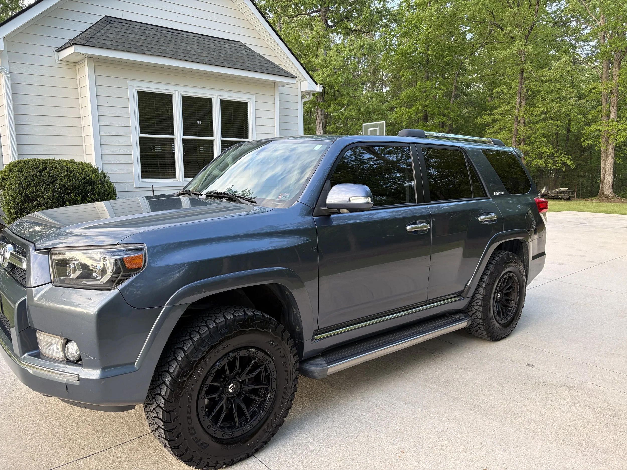 A dark gray Toyota 4Runner SUV parked on a driveway in front of a white house with large windows. Green trees are in the background.