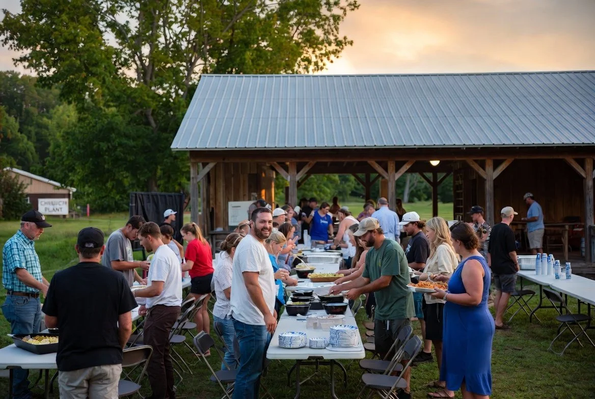 People gather at a community outdoor picnic near a wooden pavilion with food tables and people serving themselves.