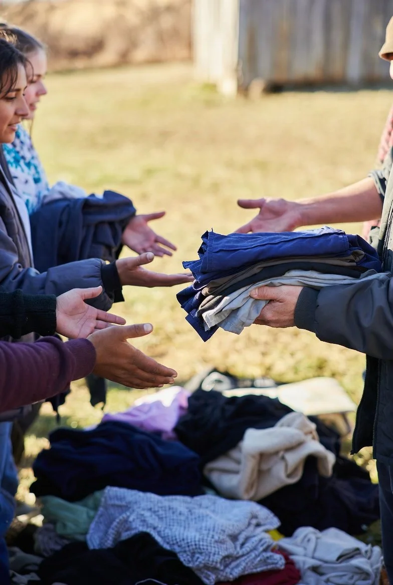 People exchanging clothes at a yard sale or donation event outdoors with a table of folded clothes in the foreground.