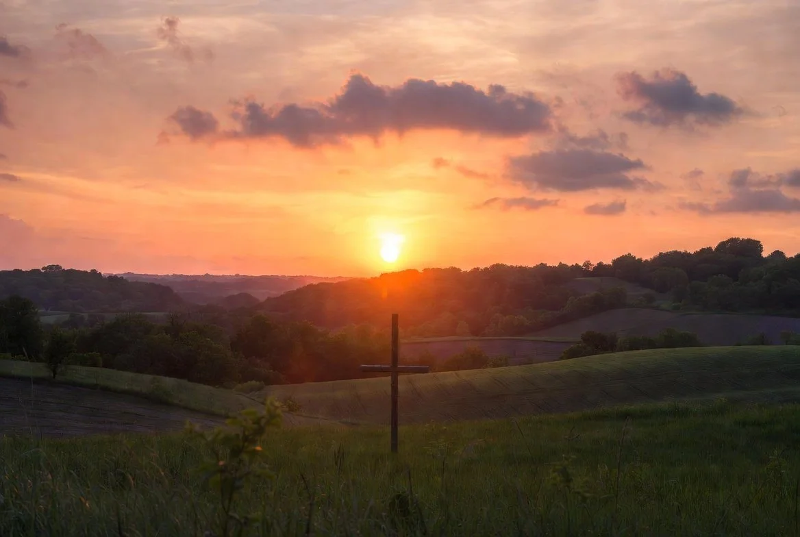 A sunset over rolling green hills with a wooden cross in the foreground and scattered clouds in the sky.