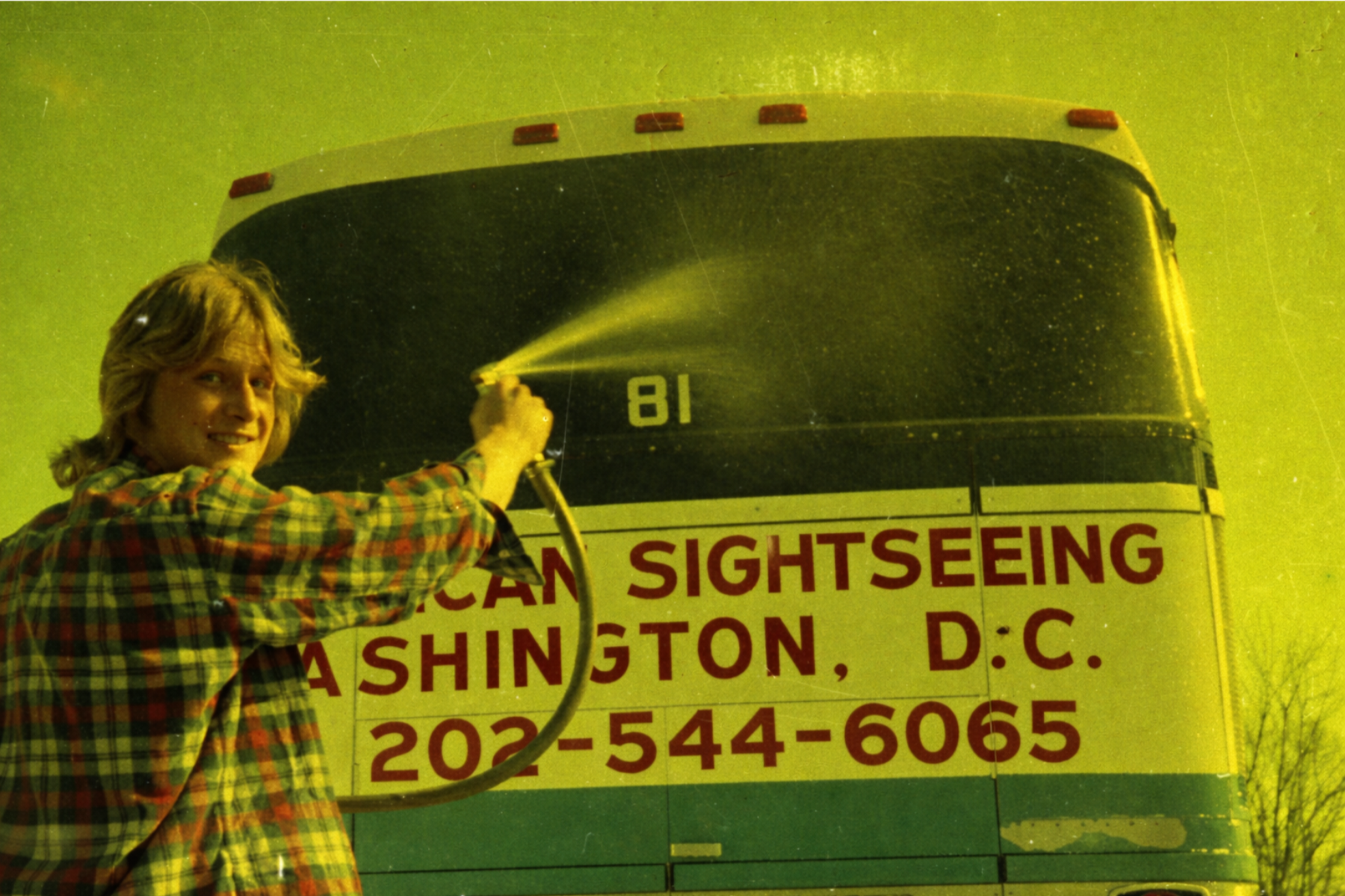 A person with long blonde hair in a plaid shirt is holding a hose and spraying water onto a bus that has a sign reading 'American Sightseeing Washington, D.C., 202-544-6065.' The bus is large, yellow, and green.