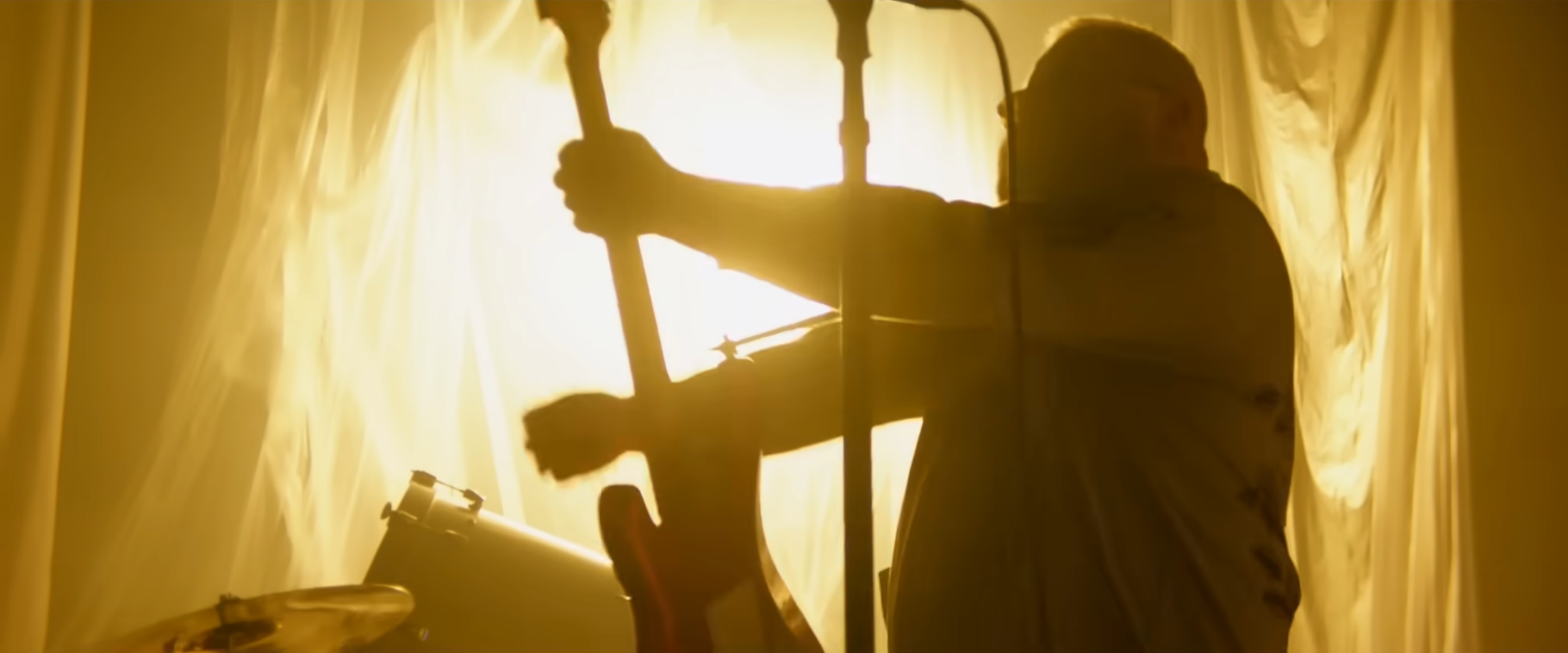 Silhouette of drummer Jordan Hastings of ALEXISONFIRE, playing drums in front of bright yellow curtains, with a guitar leaning against furniture nearby. Music video Sweet Dreams of Otherness.