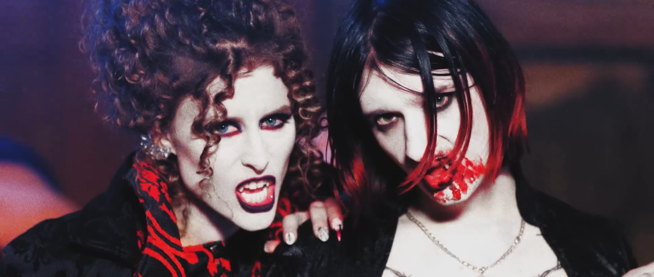 Two women with gothic makeup and dark clothing. The woman on the left has curly hair, light skin, and is growling with red lipstick. The woman on the right has straight black hair with red tips, pale skin, and red lipstick, music video KIESZA sensuum