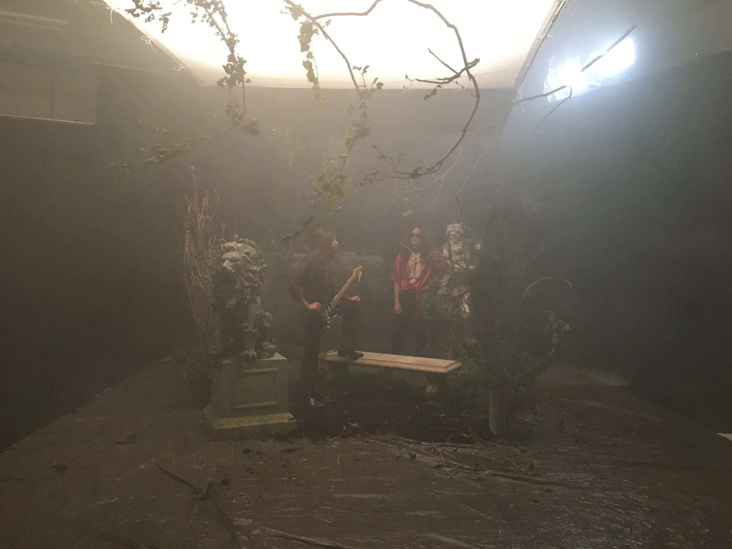 A dimly lit room filled with smoke, with two women standing near a classical sculpture of a lion and a mannequin wearing sunglasses and a red jacket. One woman is holding a guitar, and the scene appears to be a makeshift stage or display area.