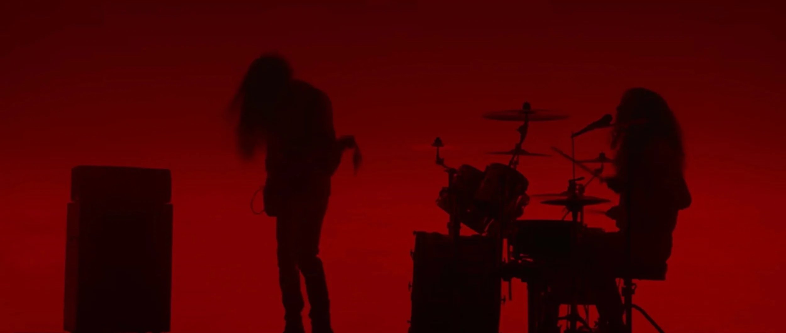 Silhouettes of two musicians from Crown Lands, with long hair playing instruments against a red background, one standing with a microphone and the other seated behind a drum set. Music video Leadfoot by Crown Lands