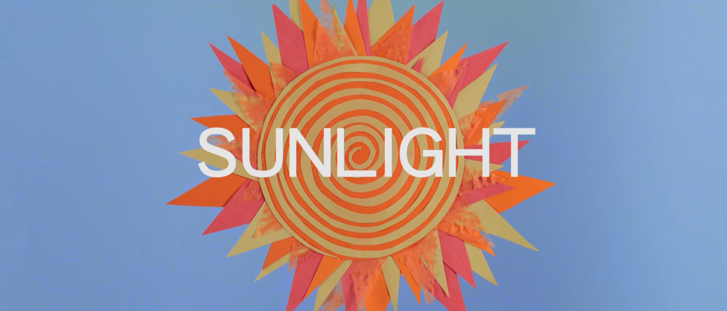 A colorful sun decoration with orange and yellow paper rays and a spiral pattern center, against a clear blue sky, with the word 'SUNLIGHT' written across it.
