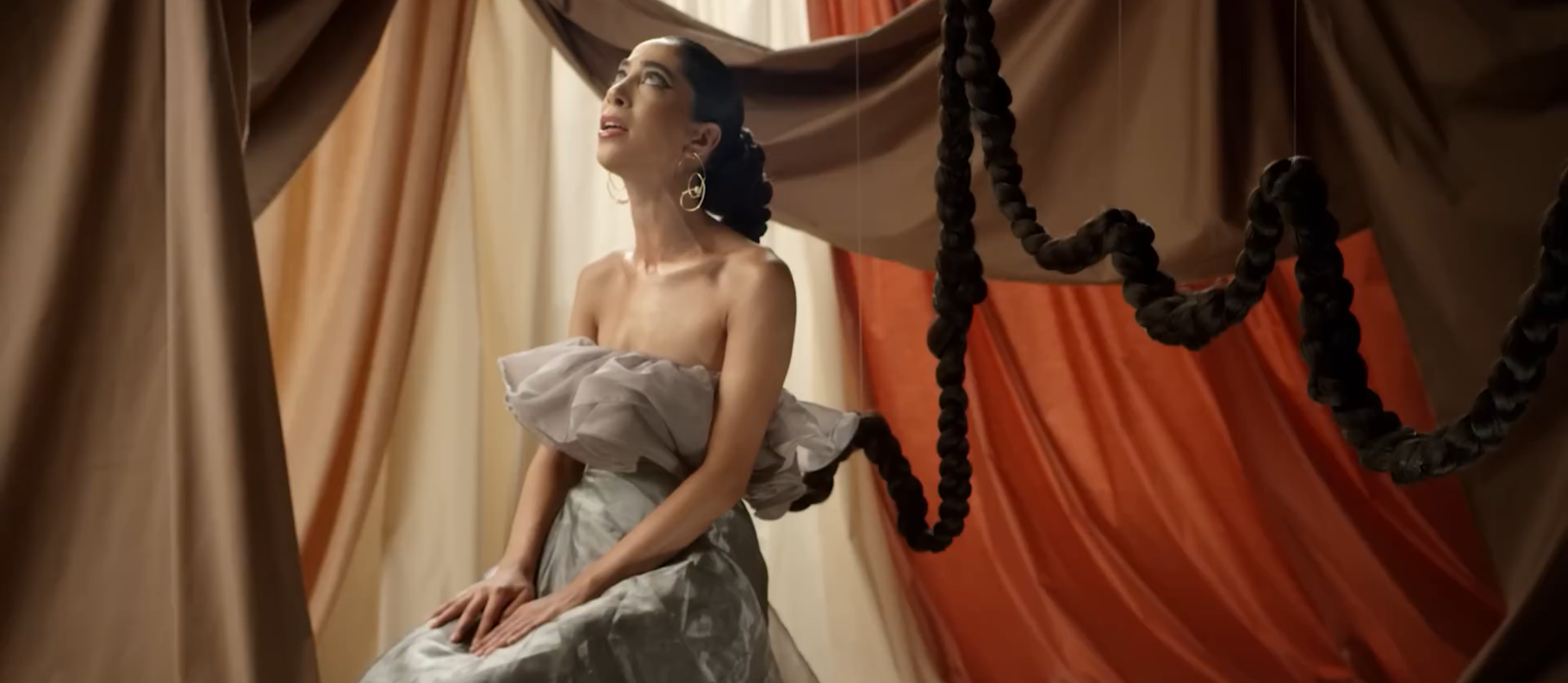 A woman with dark hair styled in braids, wearing hoop earrings, is sitting topless on a beige fabric-covered surface, with large draped curtains of beige, red, and brown in the background.