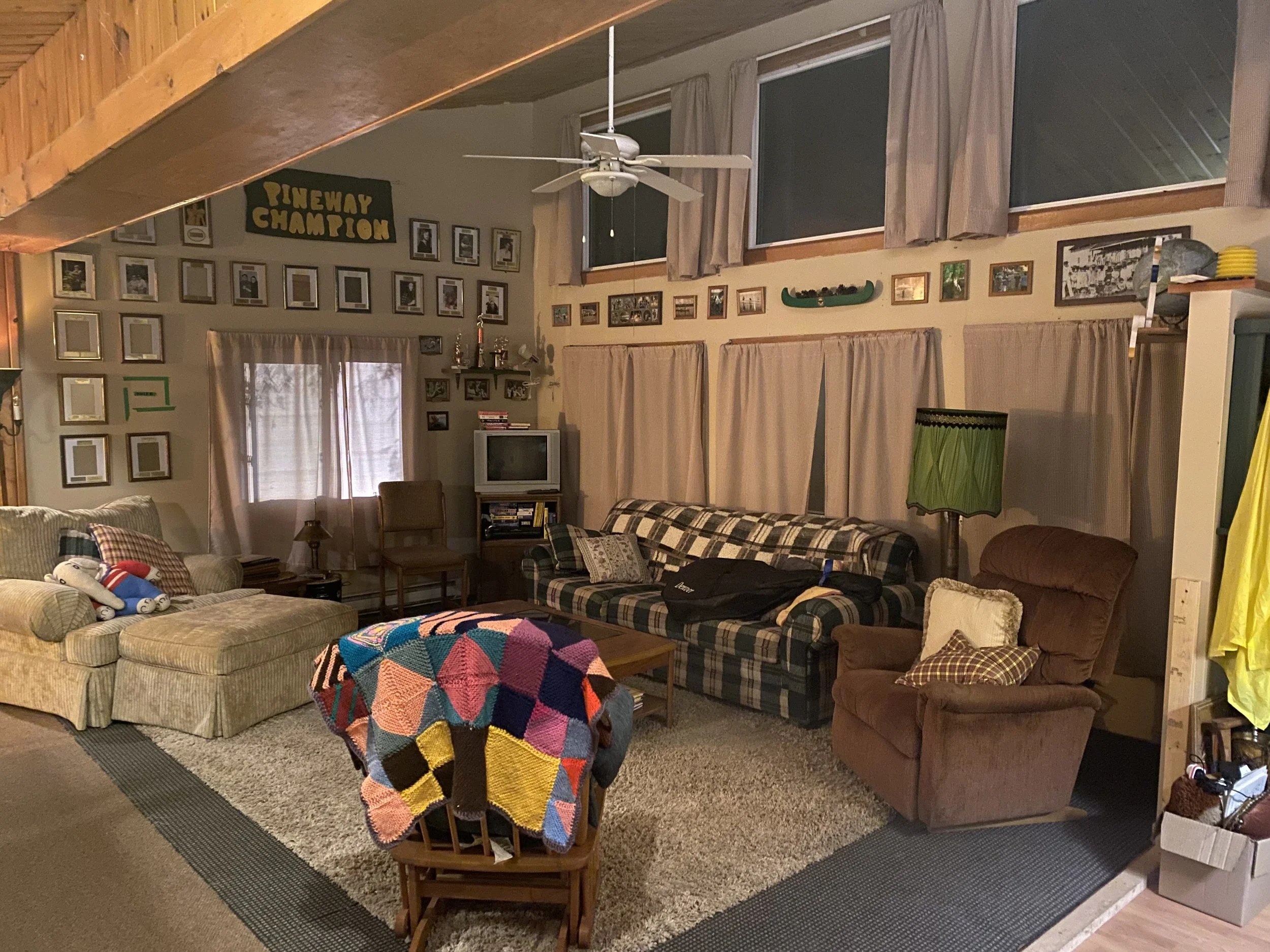 Cozy living room with sofas, a patterned armchair, a colorful crocheted blanket on a chair, framed photos, a vintage TV, and various decorative items. The walls are decorated with numerous picture frames and a sign that says "PINEWAY CHAMPION."