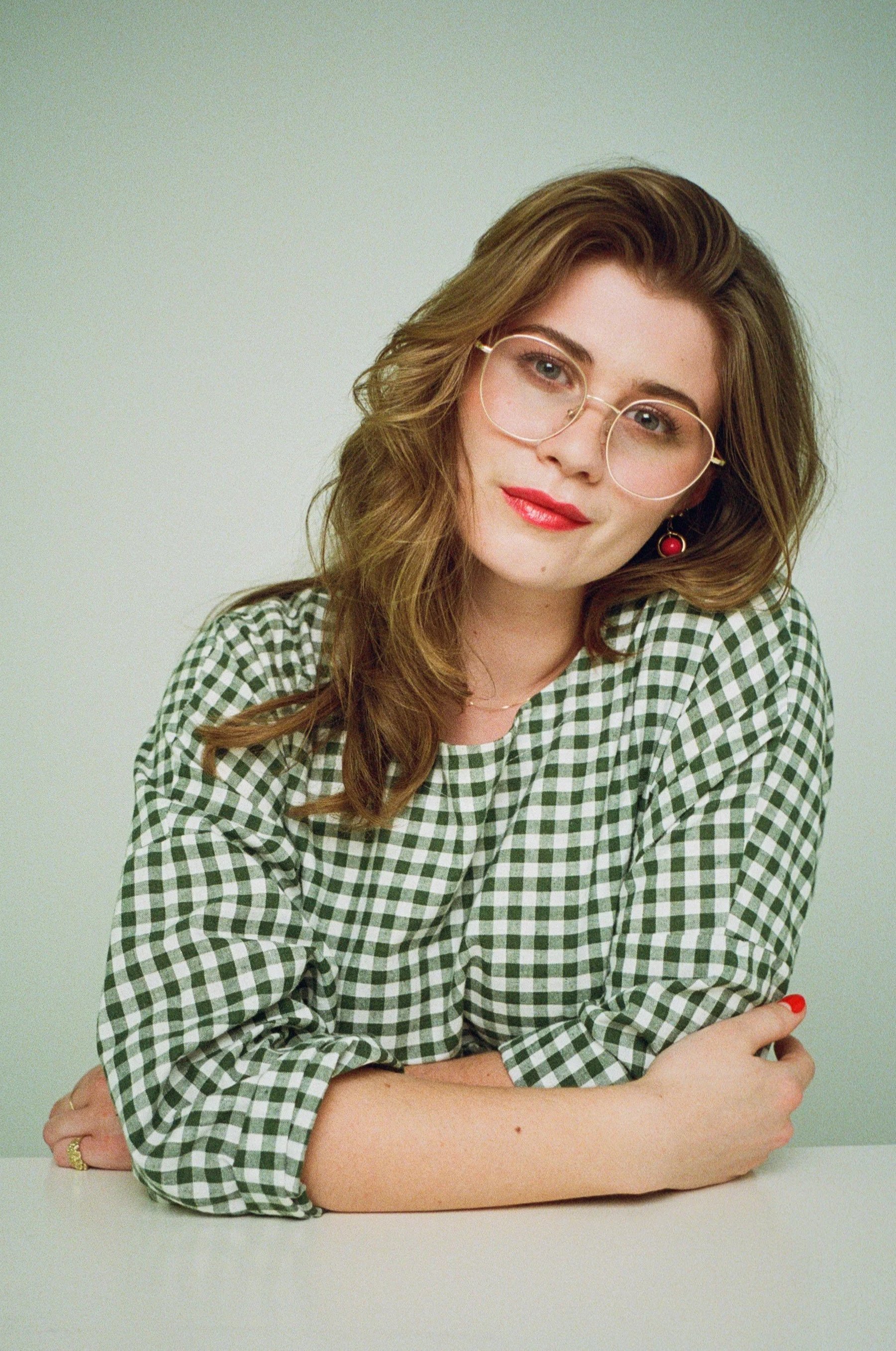 A young woman with long wavy brown hair, wearing glasses with gold frames, a green and white checkered shirt, red lipstick, red earrings, and red nail polish, sitting at a white table against a plain light-colored background. Chareese Steinhoff
