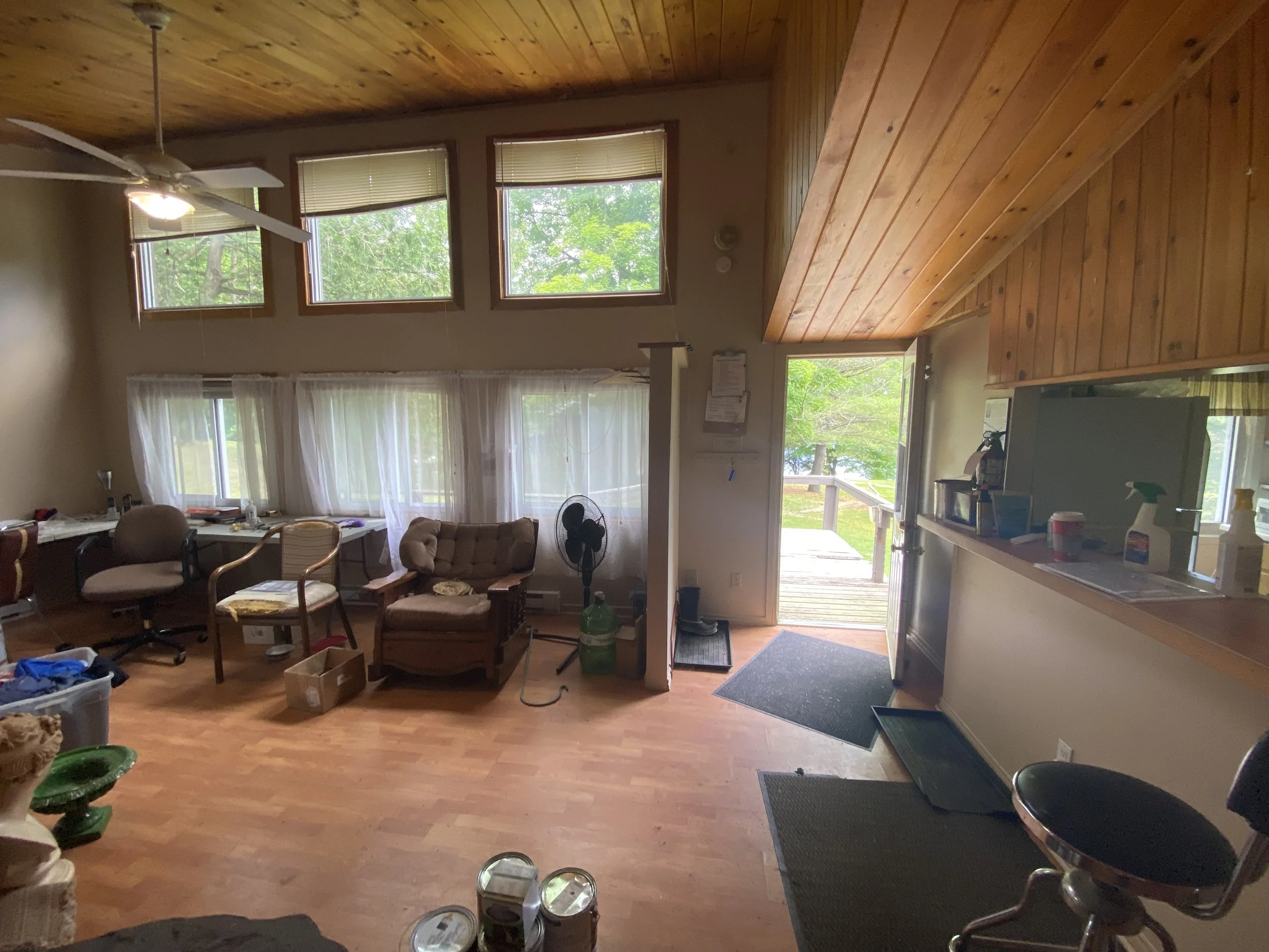 Interior of a room with wooden ceiling and walls, featuring couches, a fan, windows with curtains, and an open door leading outside to a deck with green trees visible.