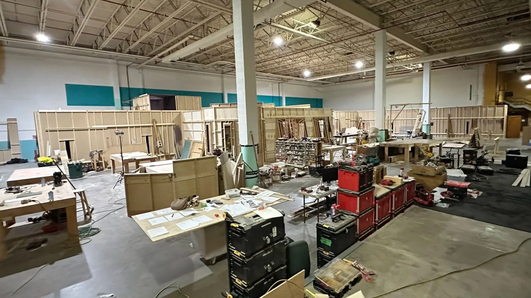 Interior of a large construction or workshop area with unfinished wooden wall panels, workbenches, tools, and equipment scattered around.