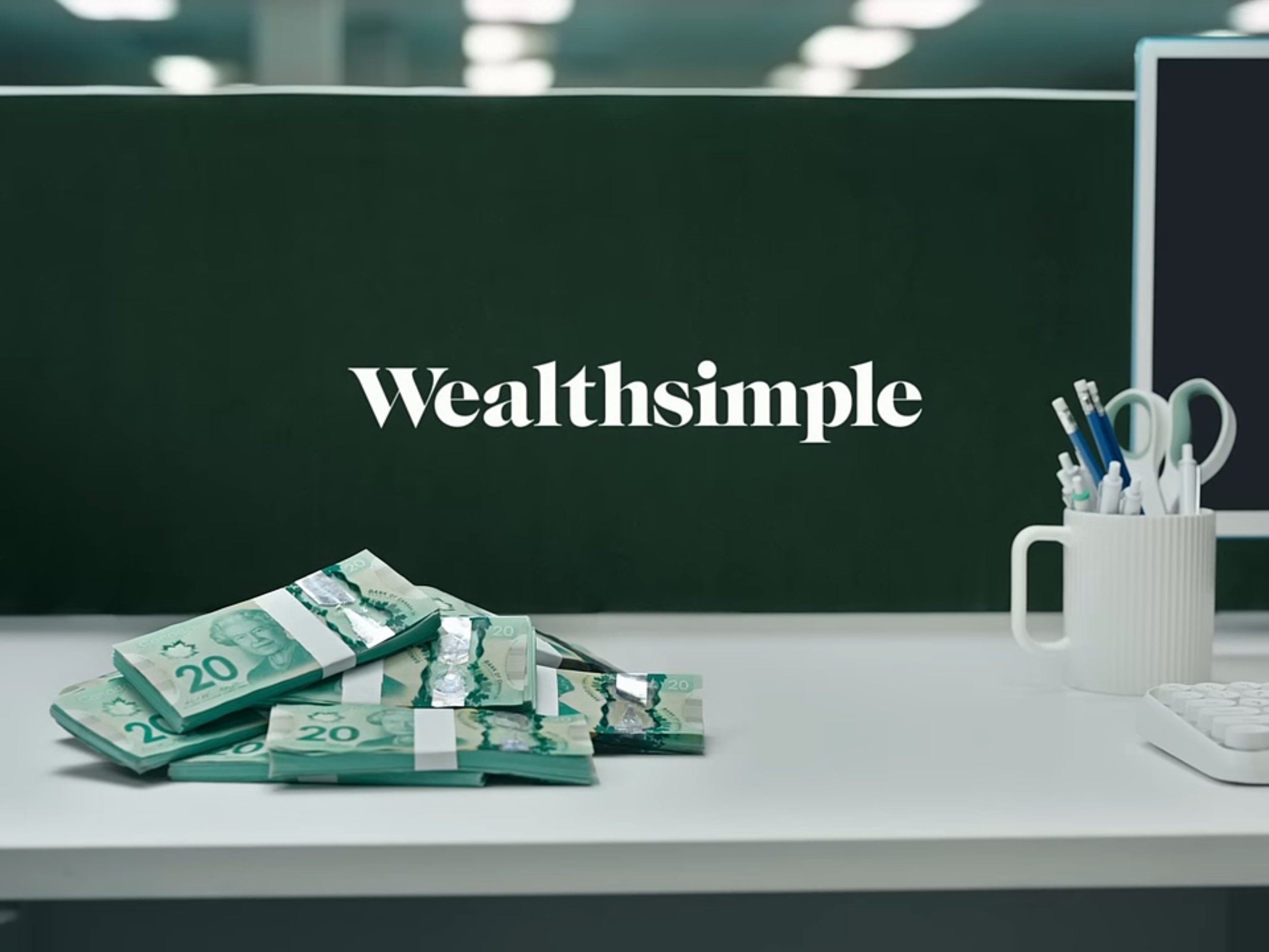 Stacks of twenty-dollar Canadian bills on a white desk, a white mug filled with pens and scissors, a computer monitor, and a blackboard with the text 'Wealthsimple' in white font.