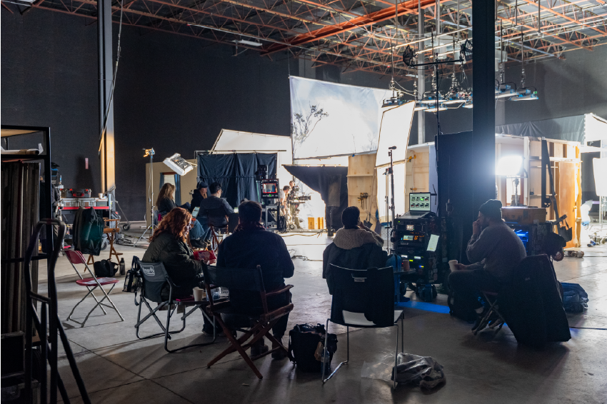 Behind the scenes of a film or commercial shoot, with crew members working on lighting, camera, and set arrangements inside a large studio space.