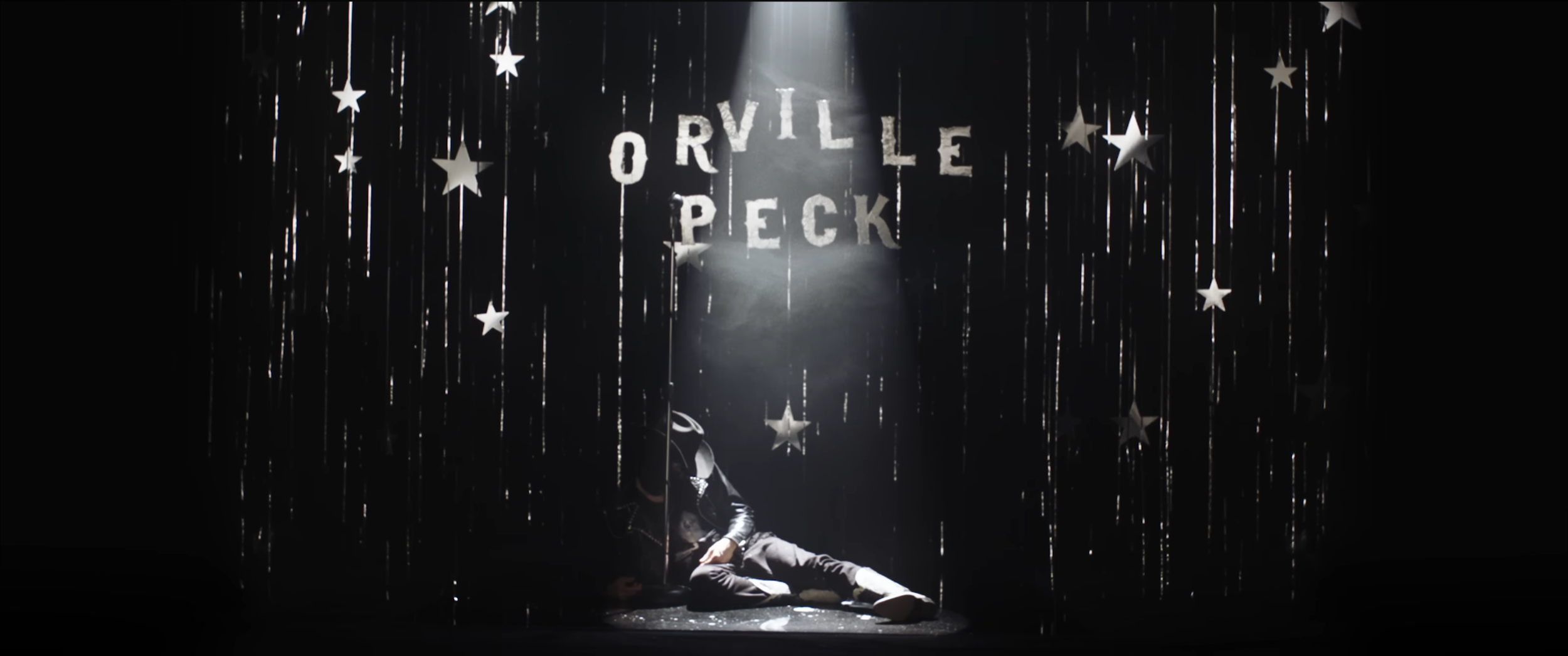 Stage decorated with hanging stars and silver streamers, with the words 'Orville Peck' displayed in large letters in the background. Music video hope to die by Orville peck