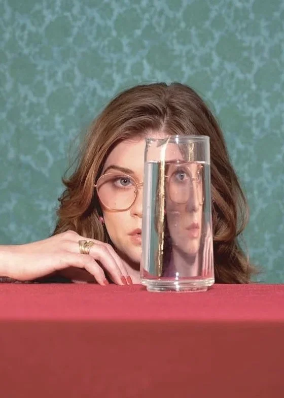 A woman with glasses and wavy brown hair looks directly into the camera, resting her hand near her face on a red surface. There is a tall glass of water placed in front of her, which creates a reflection that makes it appear as if the woman's face is doubled and distorted within the glass.