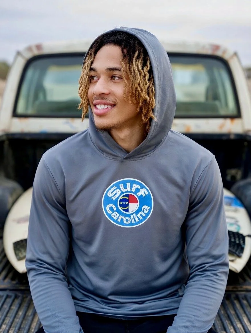 Young man with curly, blond-tipped hair wearing a gray hoodie with a circular surf Carolina logo, sitting in front of a rusty white truck with surfboards in the background.