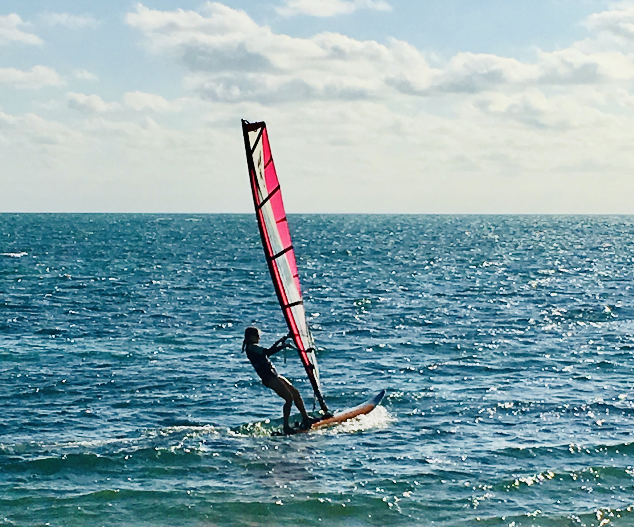 A person windsurfing on the ocean with a colorful sail under a partly cloudy sky.