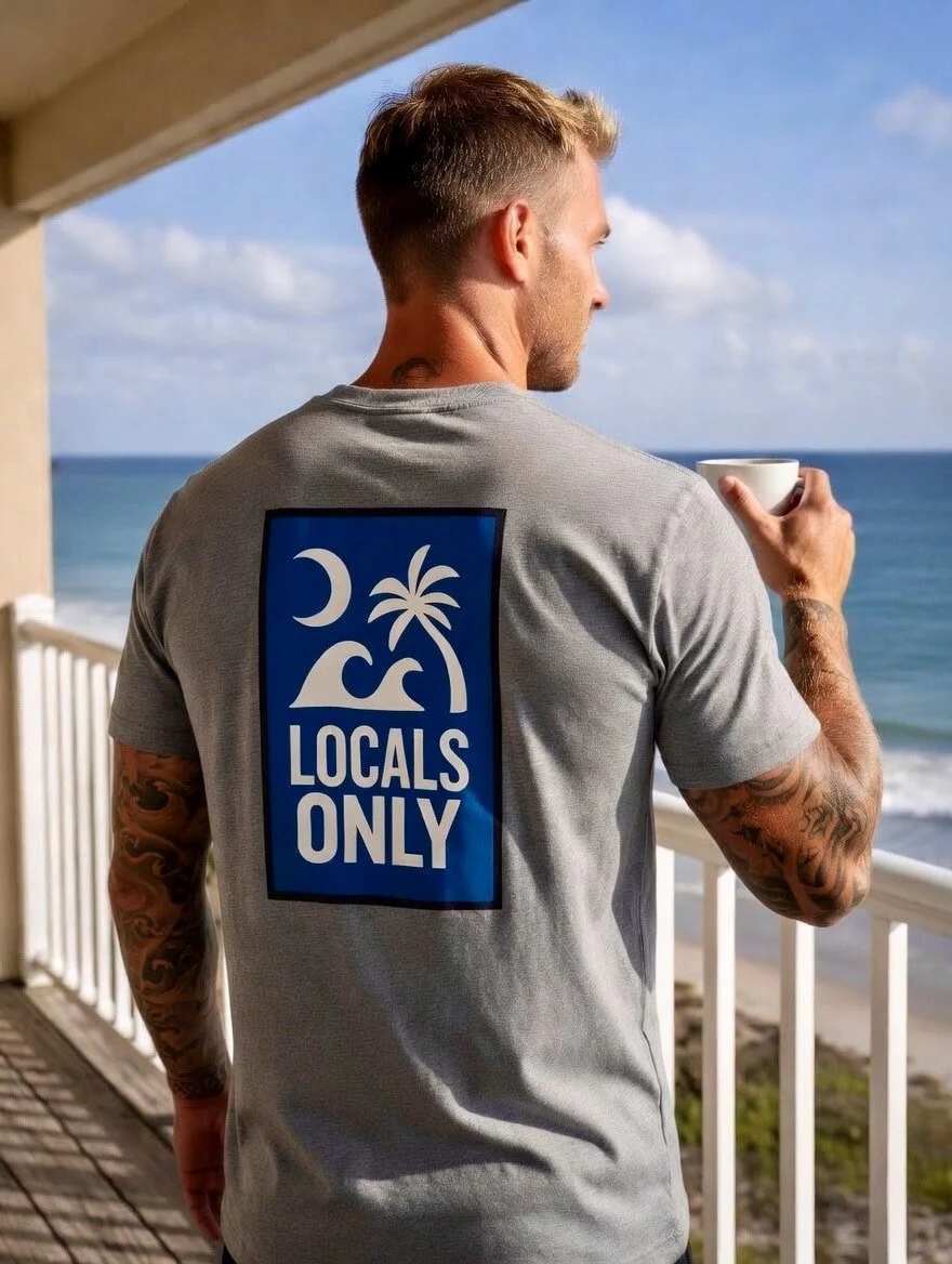 A man with tattoos on his arms is standing on a balcony overlooking the beach and ocean, holding a white mug, wearing a gray t-shirt with a 'Locals Only' logo featuring a moon, waves, a palm tree, and a mountain.