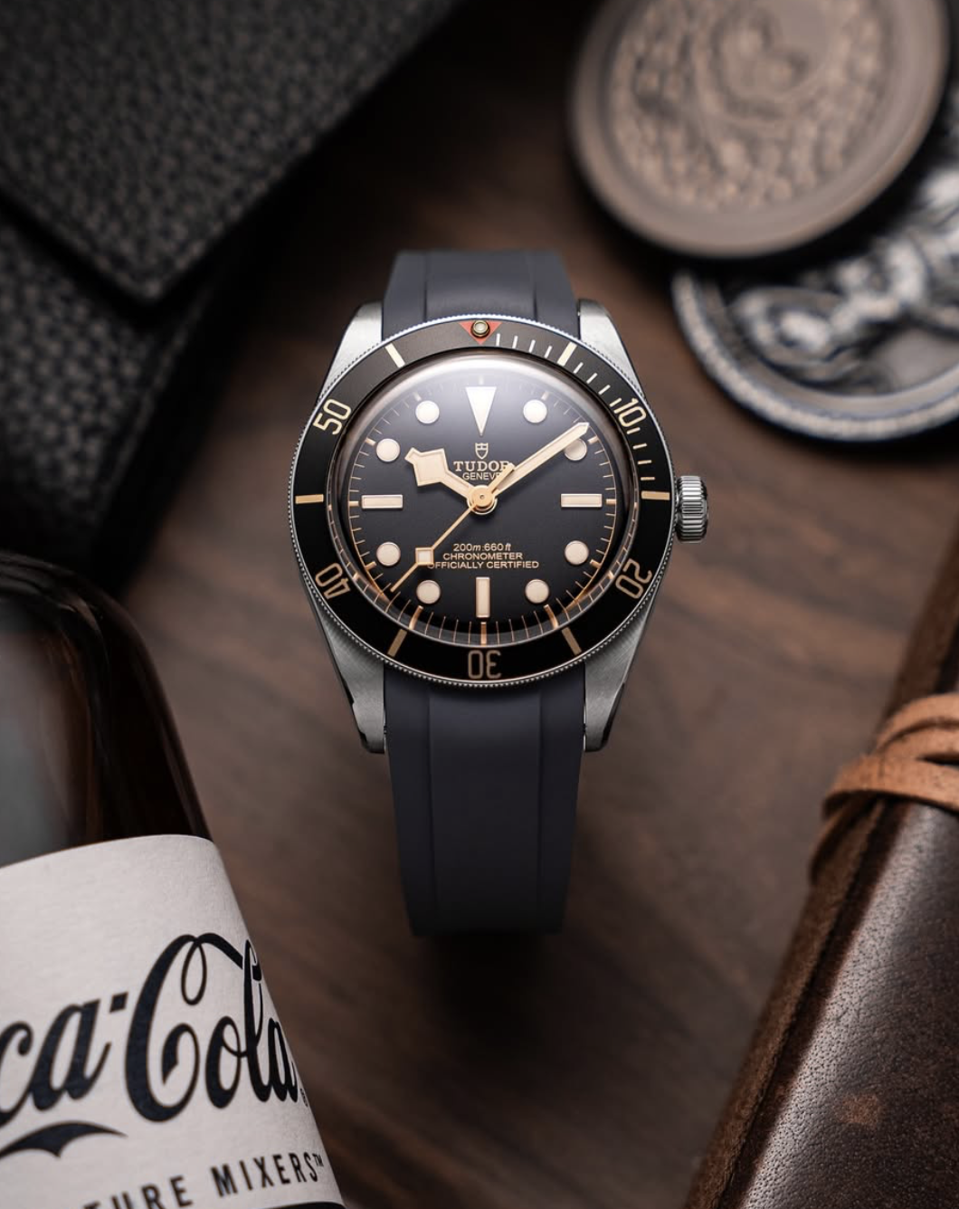 A Tudor dive watch with a black bezel and dial, gold accents, and a black strap, placed on a wooden surface among various accessories.